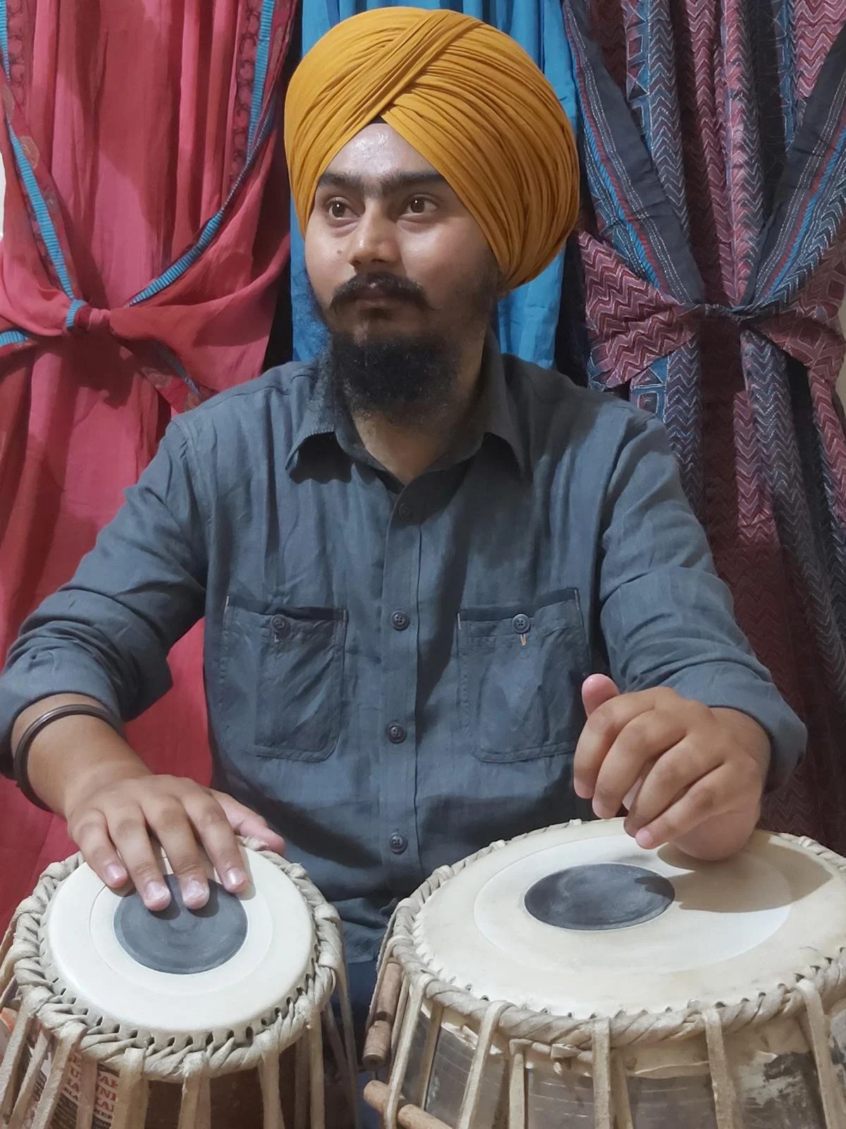 A man wearing a yellow turban and a gray shirt is playing two traditional drums in a room with colorful curtains in the background.