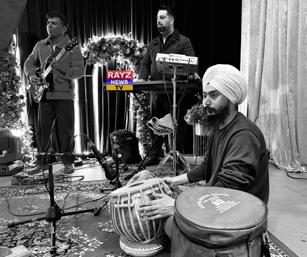 Three musicians performing on stage, with one playing an electric guitar, one on electronic equipment, and one seated on the floor playing a tabla, with microphones and floral decorations around.