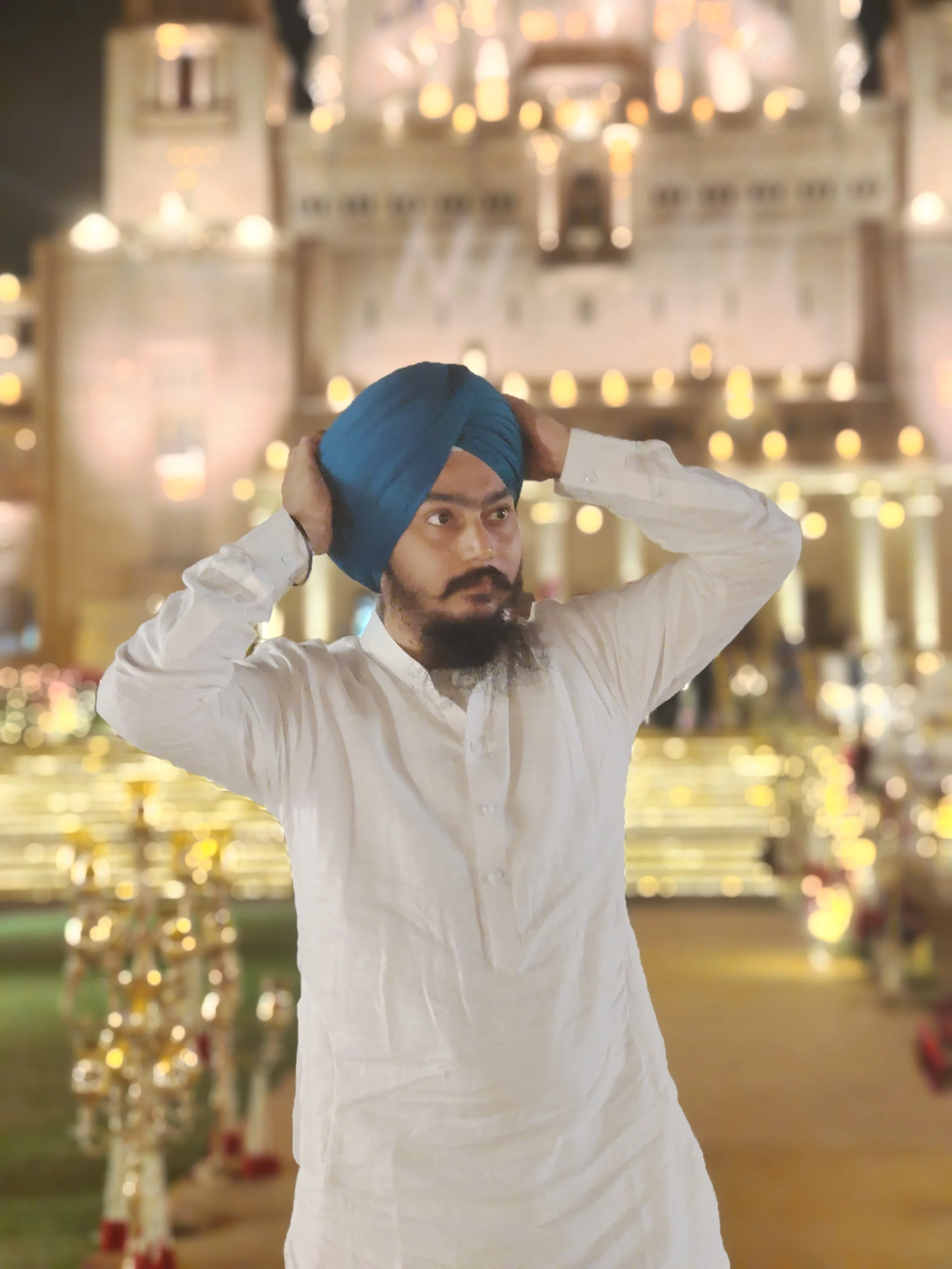 A man wearing a white kurta and a blue turban adjusting his turban in front of a decorated background with warm lights.