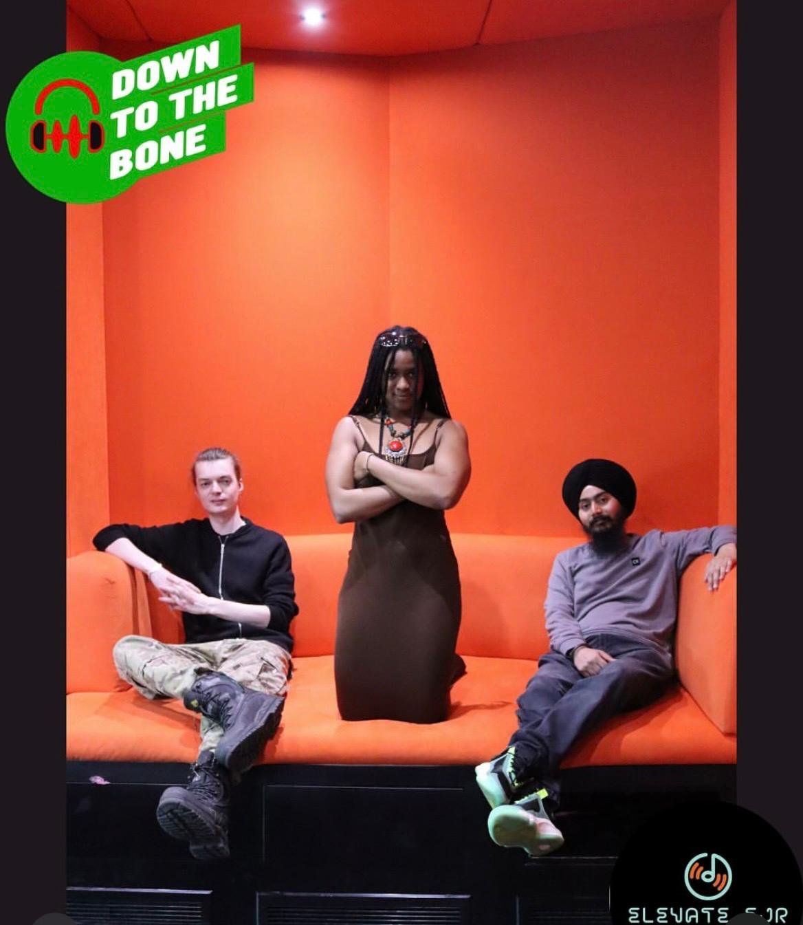 Three people sitting and standing on an orange couch in front of an orange wall, with a sticker that reads "Down to the Bone."