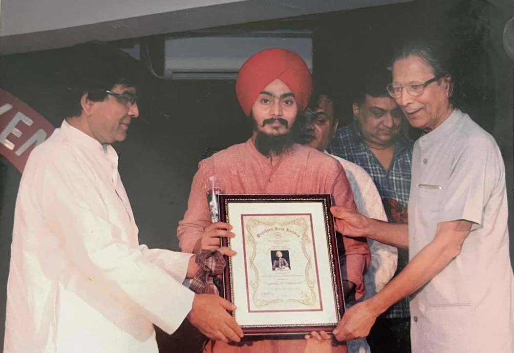 A man wearing traditional Indian attire with a red turban and beard receives a framed certificate from an older man wearing glasses and a white kurta. Two other men watch and stand nearby in a formal setting.