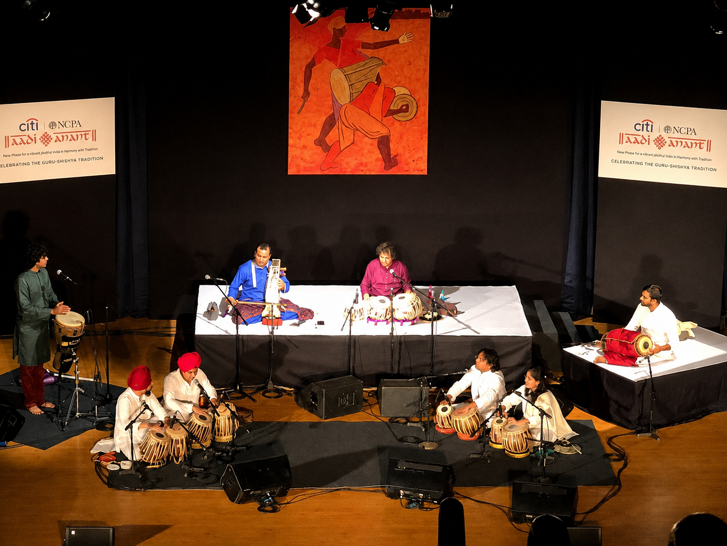 A group of musicians performing on a stage with traditional Indian instruments, including tabla drums and other percussion, with two singers seated at the center. The stage has a large back curtain with a colorful painting or tapestry of a dancer, and banners on either side that read 'Celebrating the Guru-Shishya Tradition'.
