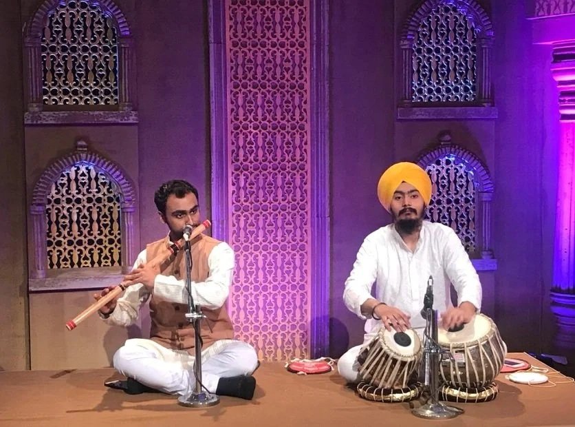 Two men are performing traditional Indian music on stage, with one playing a flute and the other playing tabla drums, against a backdrop with ornate arches and a purple lighting scheme.