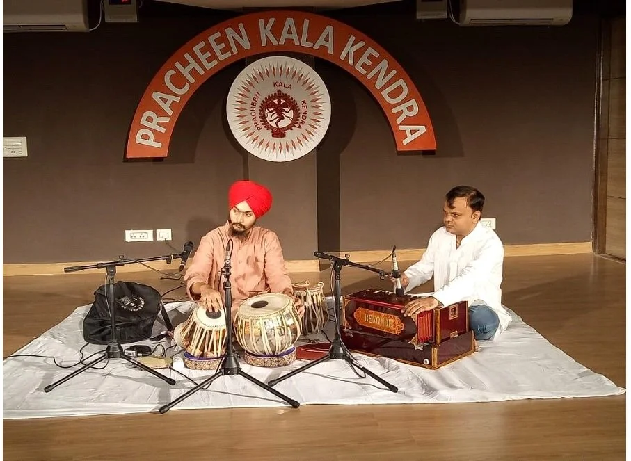 Two men sitting on a white cloth playing Indian classical music instruments in front of a red and white sign that reads "Praacheen Kala Kendra." The man on the left is wearing a red turban and beige traditional attire, playing a tabla, while the man on the right is in a white shirt, playing a harmonium.