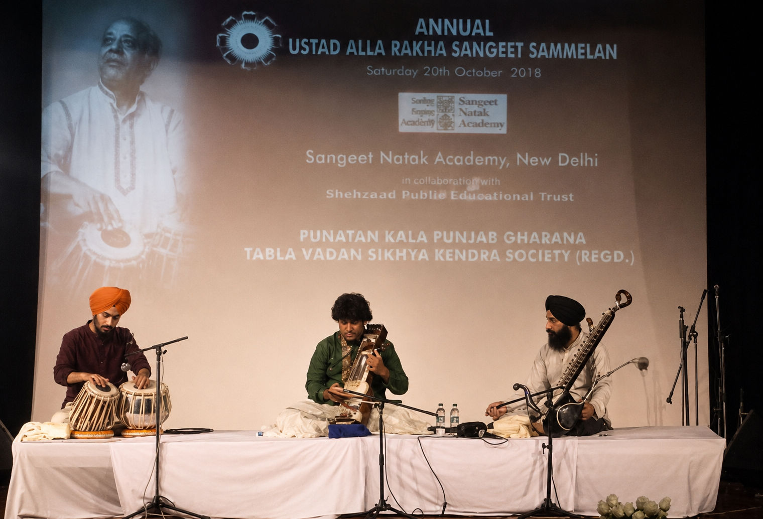 Three musicians performing on stage at the annual Ustad Alla Rakha Sangeet Sammelan. The event took place on October 20, 2018, in New Delhi. The musicians are playing traditional Indian instruments, with a large screen behind them displaying event details and logos.