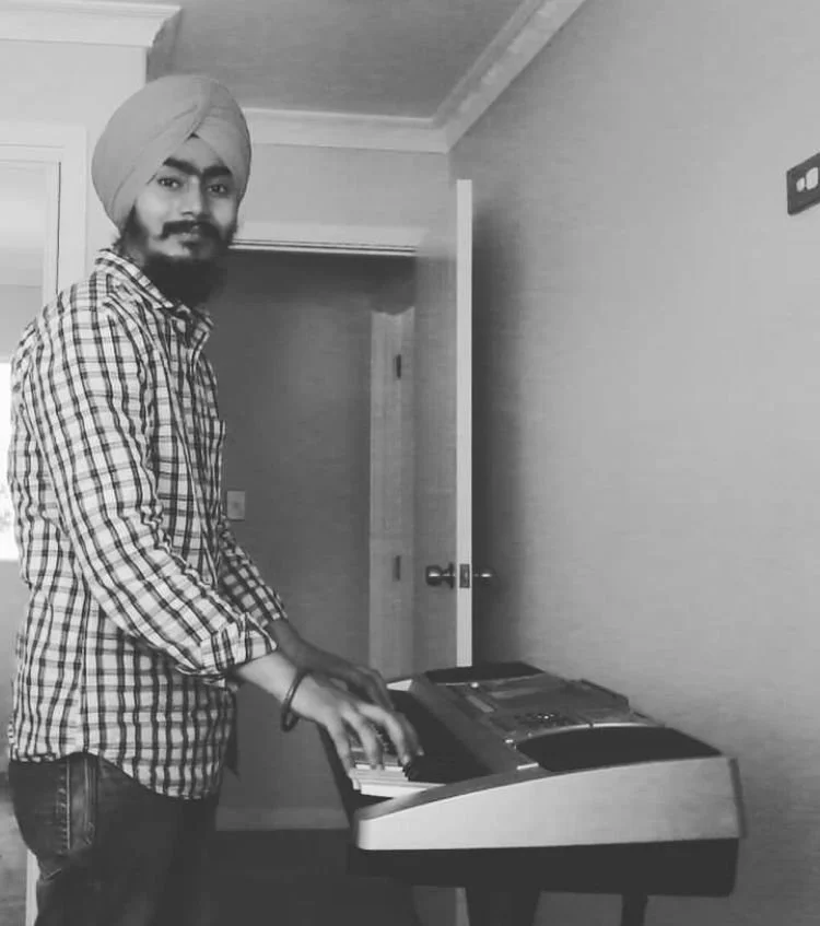 A man wearing a turban, checkered shirt, and jeans playing an electronic keyboard in a room with a door open behind him.