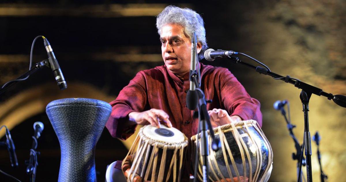 A man with gray hair wearing a red shirt playing a tabla on a stage with microphones.