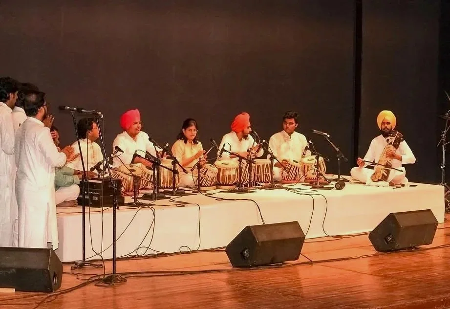 Indian classical musicians performing on stage with traditional instruments and singers, wearing white traditional attire, some with turbans.
