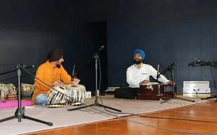 Two musicians sitting on a stage, one playing tabla drums and the other playing harmonium, with microphones set up in front of them.