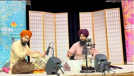 Two men wearing traditional Punjabi turbans sitting on a stage, playing musical instruments with a yellow and a purple room divider in the background.