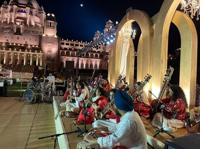 Musicians dressed in traditional attire performing on an outdoor stage at night with a palace in the background and illuminated arches behind them.