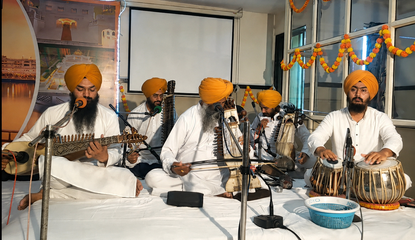 Group of Sikh men dressed in white traditional attire and orange turbans performing Indian classical music. They are playing various instruments including a harmonium, tabla, and stringed instrument, seated on a white stage decorated with orange marigold garlands.