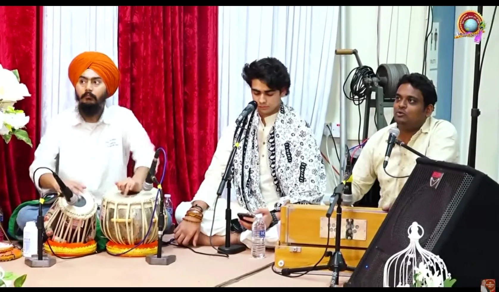 Three men performing traditional Indian music, with two playing tabla drums and one with a harmonium, on a stage with red curtains.