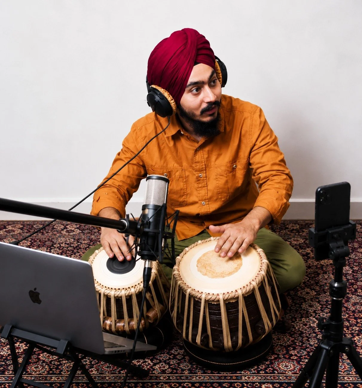 A man wearing a red turban and an orange shirt, sitting on a patterned rug, plays traditional Indian drums called tabla while recording a music session with a laptop, microphone, headphones, and a smartphone.
