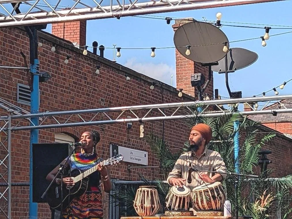 A woman singing and playing guitar, and a man playing drums on a small outdoor stage with string lights and tropical plants.
