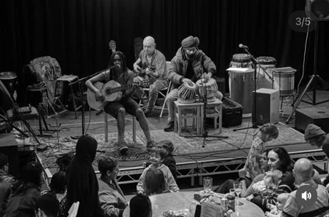 Musicians performing on stage with a guitarist, percussionist, and other players, while an audience watches in a seated area.