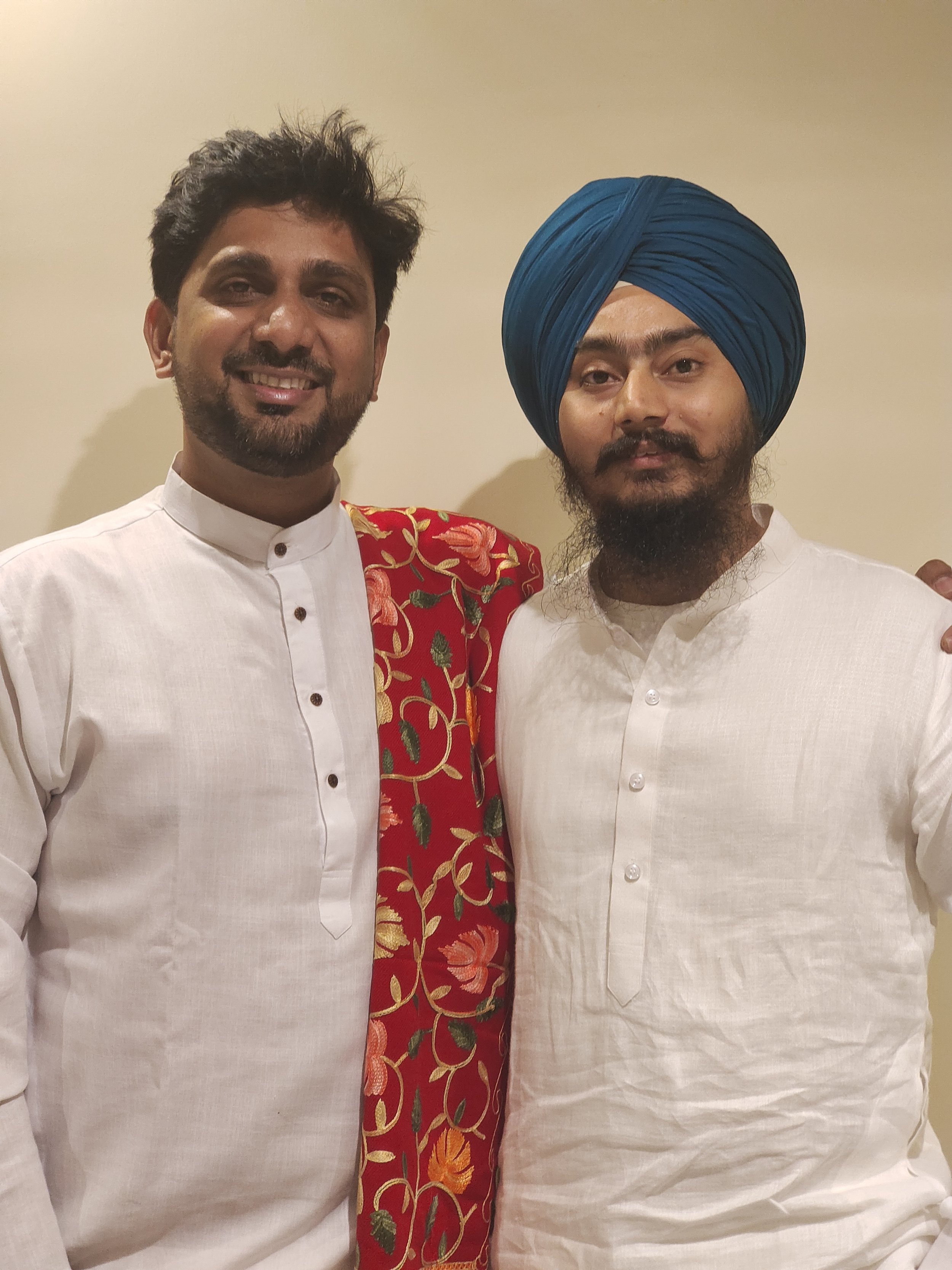 Two men dressed in traditional Indian attire, one wearing a white kurta and the other wearing a cream kurta with a blue turban, standing side by side indoors against a plain wall.