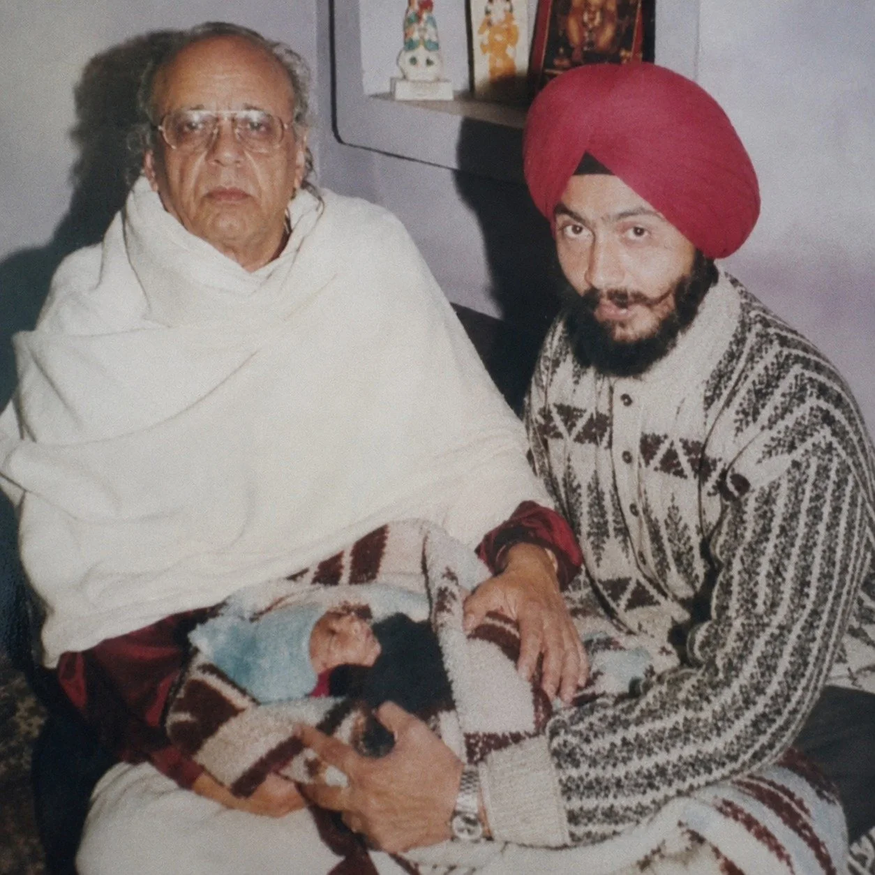 An elderly man dressed in white and an older man wearing a red turban are sitting together, holding a newborn baby wrapped in blankets. The background shows a wall with religious or cultural items and framed pictures.