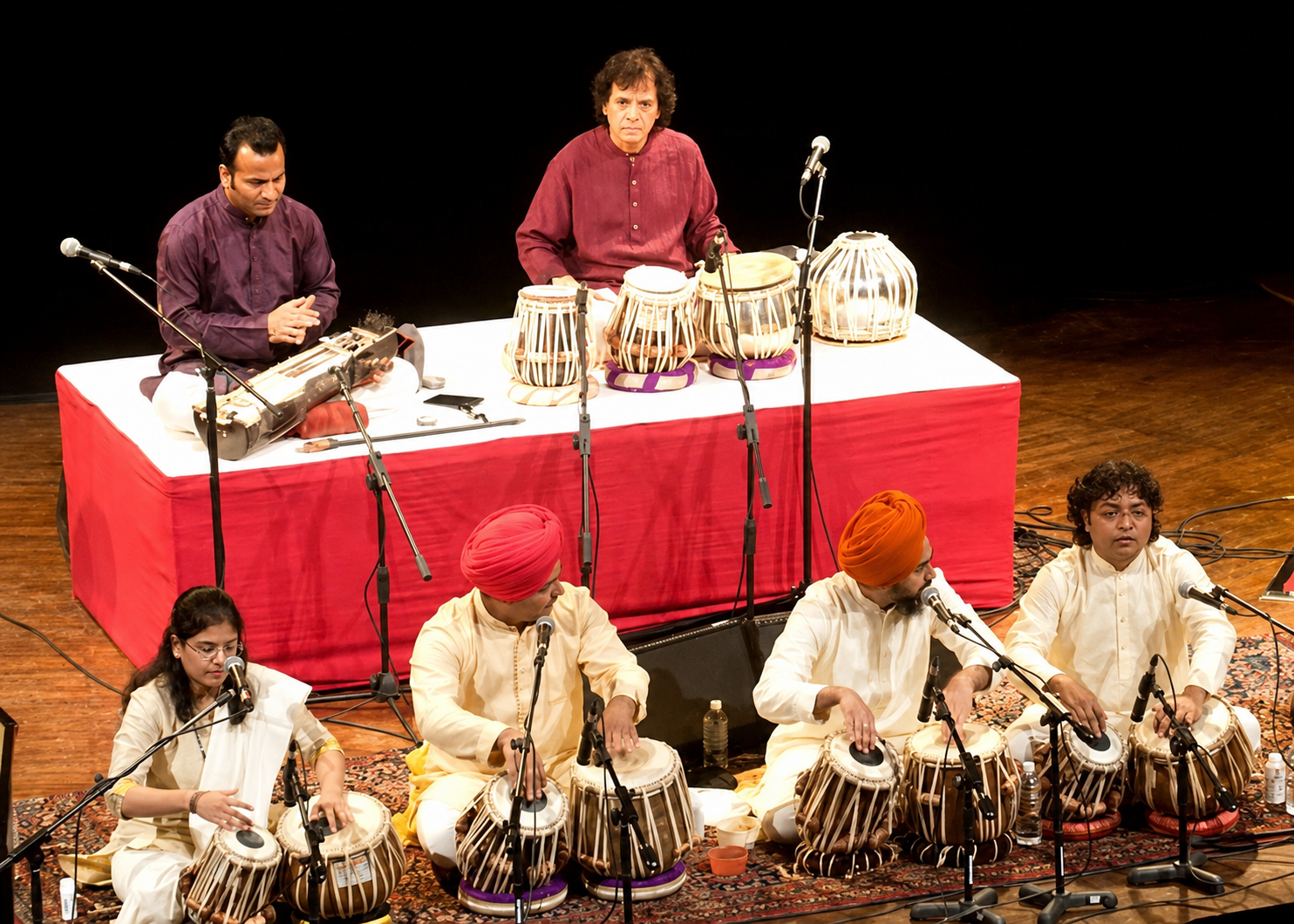 Musicians performing Indian classical music on stage, with four musicians seated and playing tabla drums, a vocalist with a harmonium, and a percussionist with drums, all dressed in traditional Indian attire.