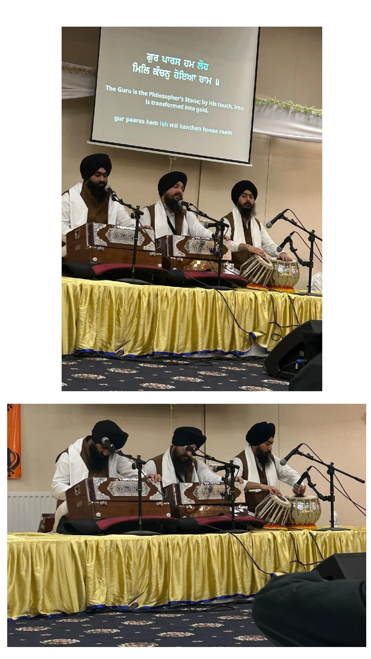 Three men wearing black turbans and traditional white and brown clothing sit on a stage, playing harmonium and tabla. Behind them is a large projection screen with text in Gurmukhi script and English, reading 'The Guru is the Philosopher's Stone; by 