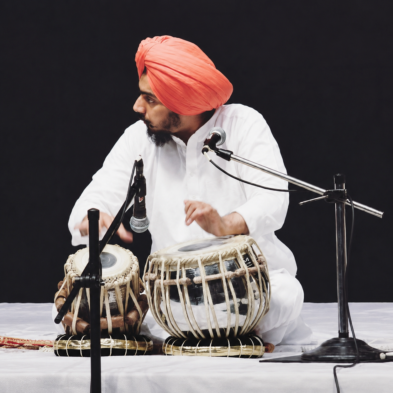 A man wearing a red turban and white traditional attire playing Indian tabla drums on stage with microphones set up.
