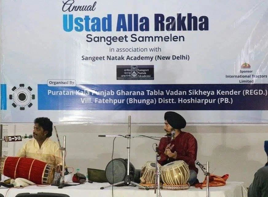 Two men sitting on a stage, playing traditional Indian musical instruments during a cultural event. A large banner behind them displays event details in English and Hindi.