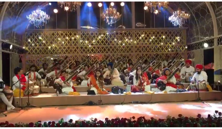 Group of musicians performing on stage with traditional Indian instruments, decorated with chandeliers and floral arrangements.