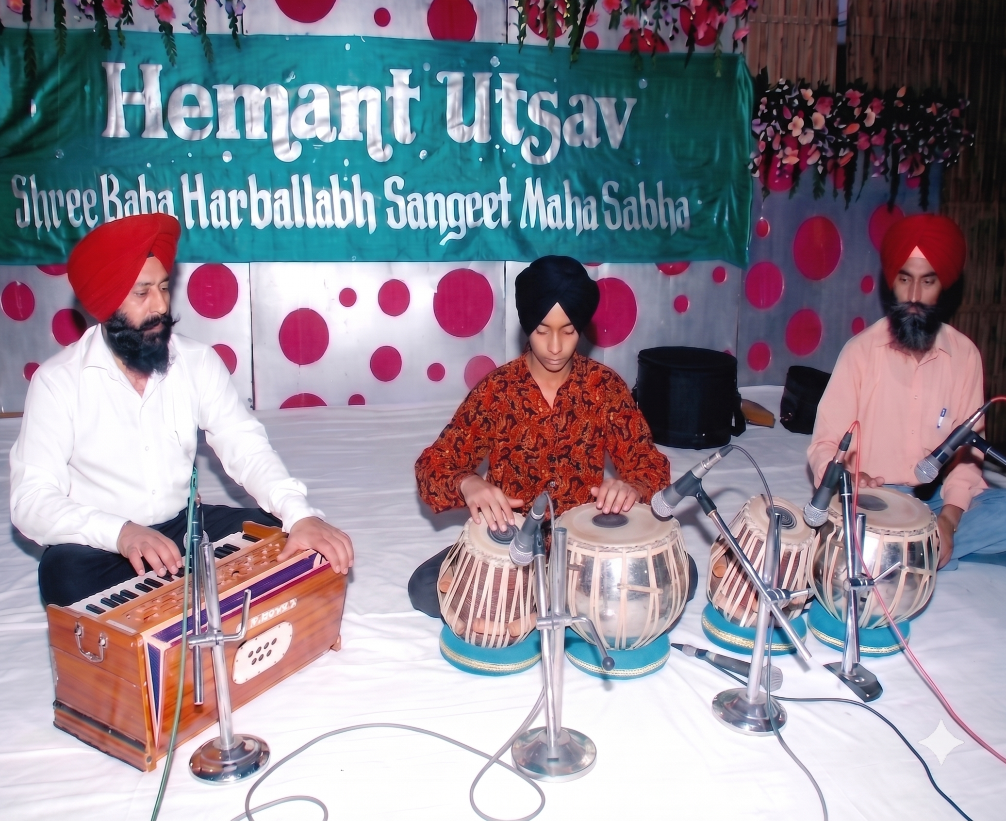 Three men in traditional Sikh turbans sitting at a table with musical instruments, including a harmonium, tabla, and keyboard, during a cultural event called Hemant Utsav, with a decorative backdrop and a banner in bright colors.