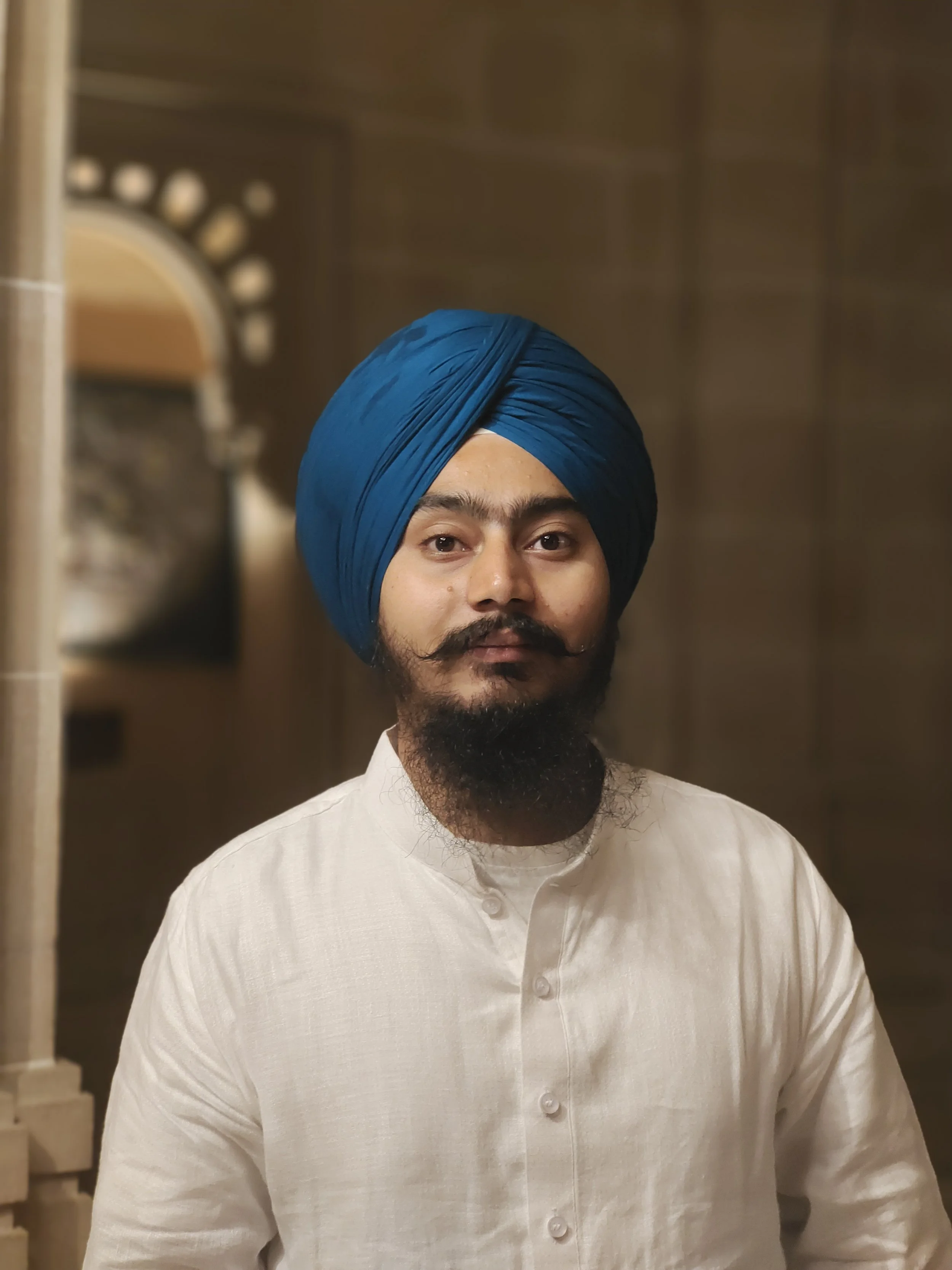 Portrait of a man wearing a blue turban and a white kurta, standing indoors against a wooden background.
