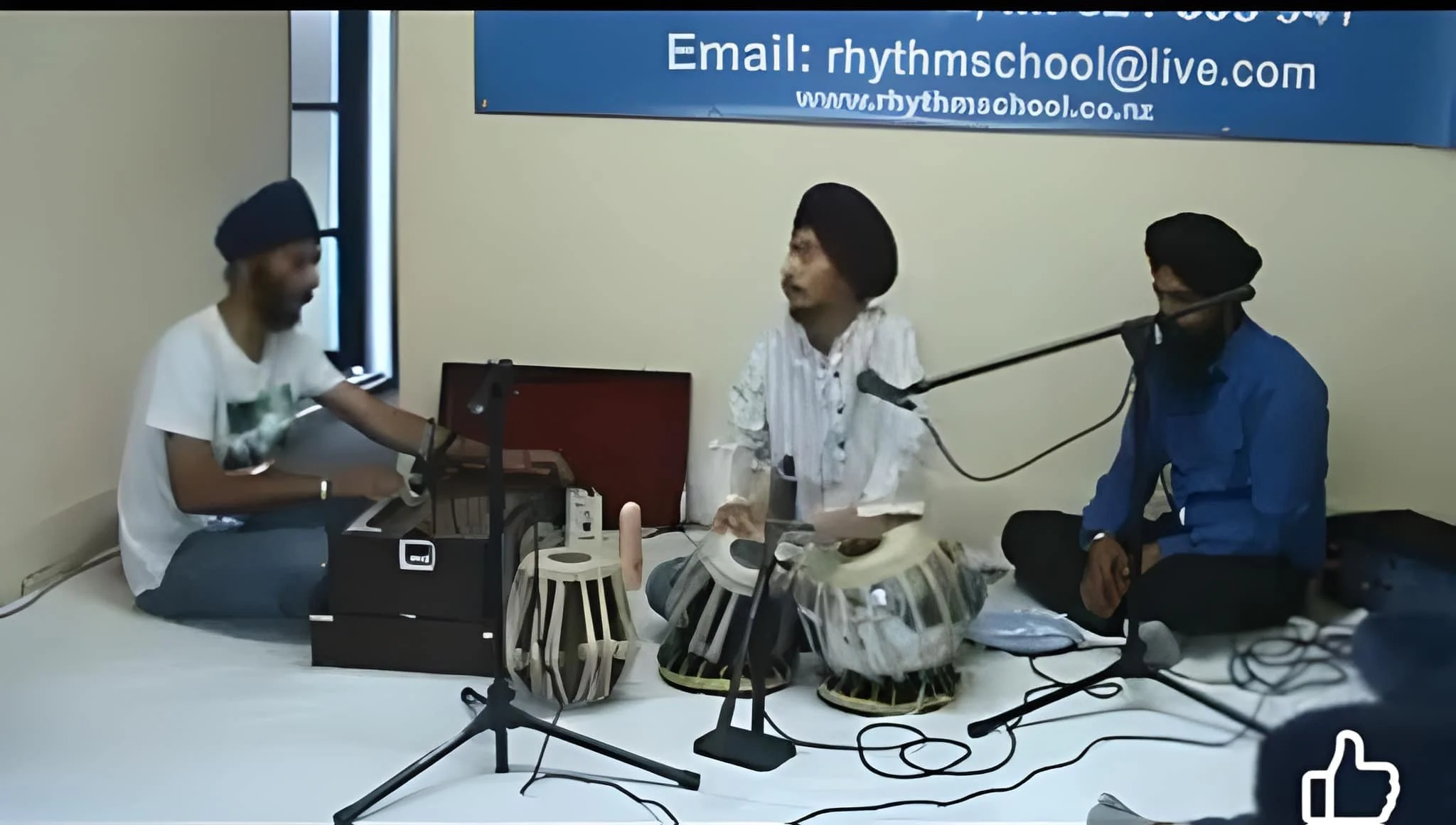 Three men in a room appear to be engaged in a music or sound experiment. One man on the left is sitting at a table with electronic sound equipment; he has a beard and is wearing a black turban and white T-shirt. The man in the middle, wearing a white