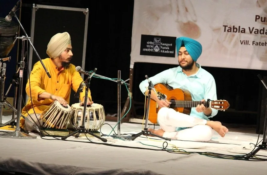 Two men, both wearing traditional turbans, are sitting on a stage. The man on the left is playing a tabla, and the man on the right is playing an acoustic guitar. They are performing together at an event, with microphones set up in front of them.