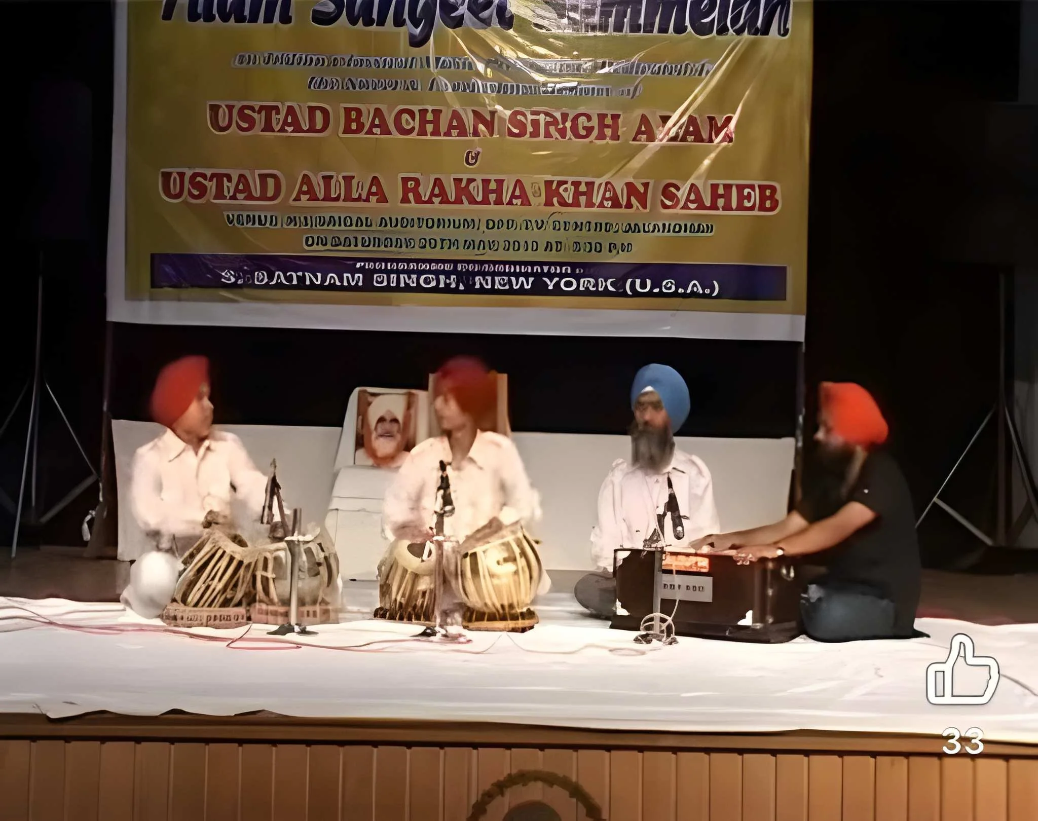 A stage with four men wearing white shirts and orange or blue turbans, performing traditional Indian drums called tabla, with a large yellow banner in the background that has text in multiple languages, including English and Hindi, indicating a cultu