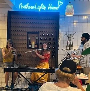 A musical performance at a cozy indoor venue with two women singing and playing instruments, a man playing a drum, a keyboard, and an audience member watching, decorated with wall art, a sign that says 'Northern Lights Home,' and hanging lights.