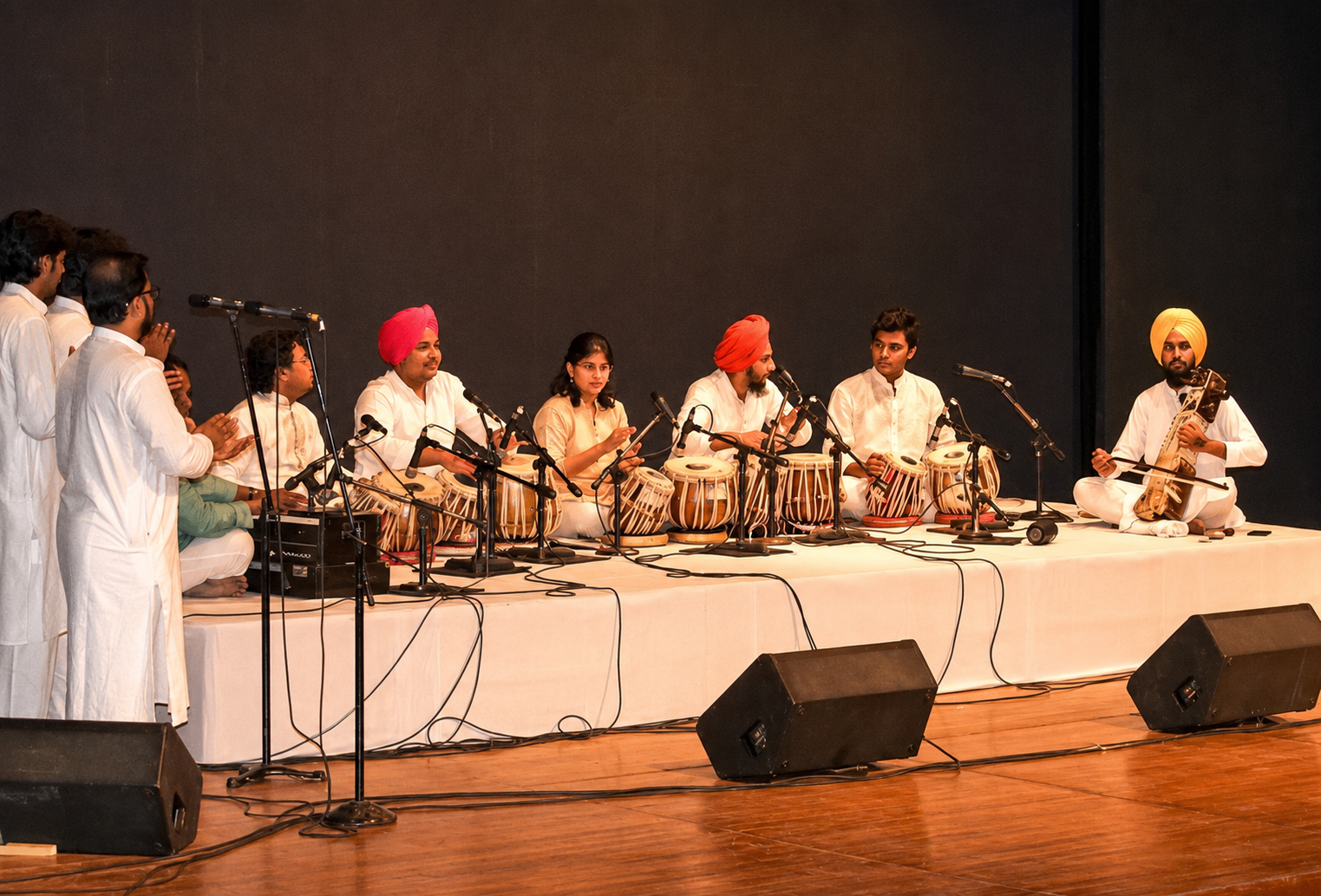 A group of musicians performing on stage with traditional Indian instruments, including tabla, guitar, and microphones. They are dressed in traditional Indian attire, with some wearing turbans, and are seated on a white stage with a black backdrop.