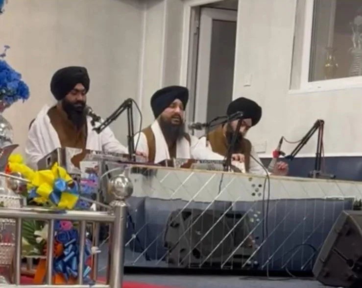 Three men wearing turbans singing or speaking into microphones at a table with flowers and gifts nearby.