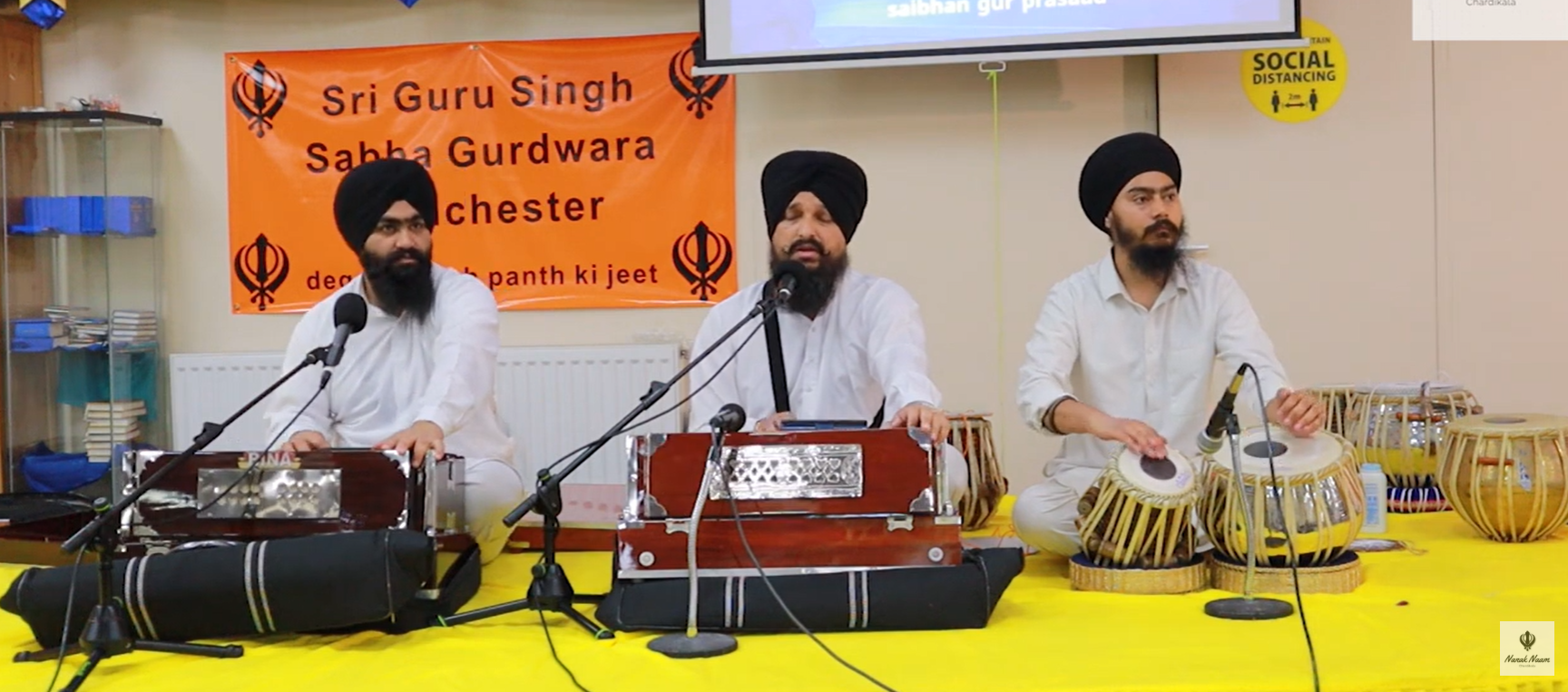 Three men wearing white traditional attire and black turbans sitting on a yellow cloth, performing Indian classical music with traditional instruments, in front of an orange banner with black text and Sikh symbols, in an indoor setting with bookshelves and social distancing signage.