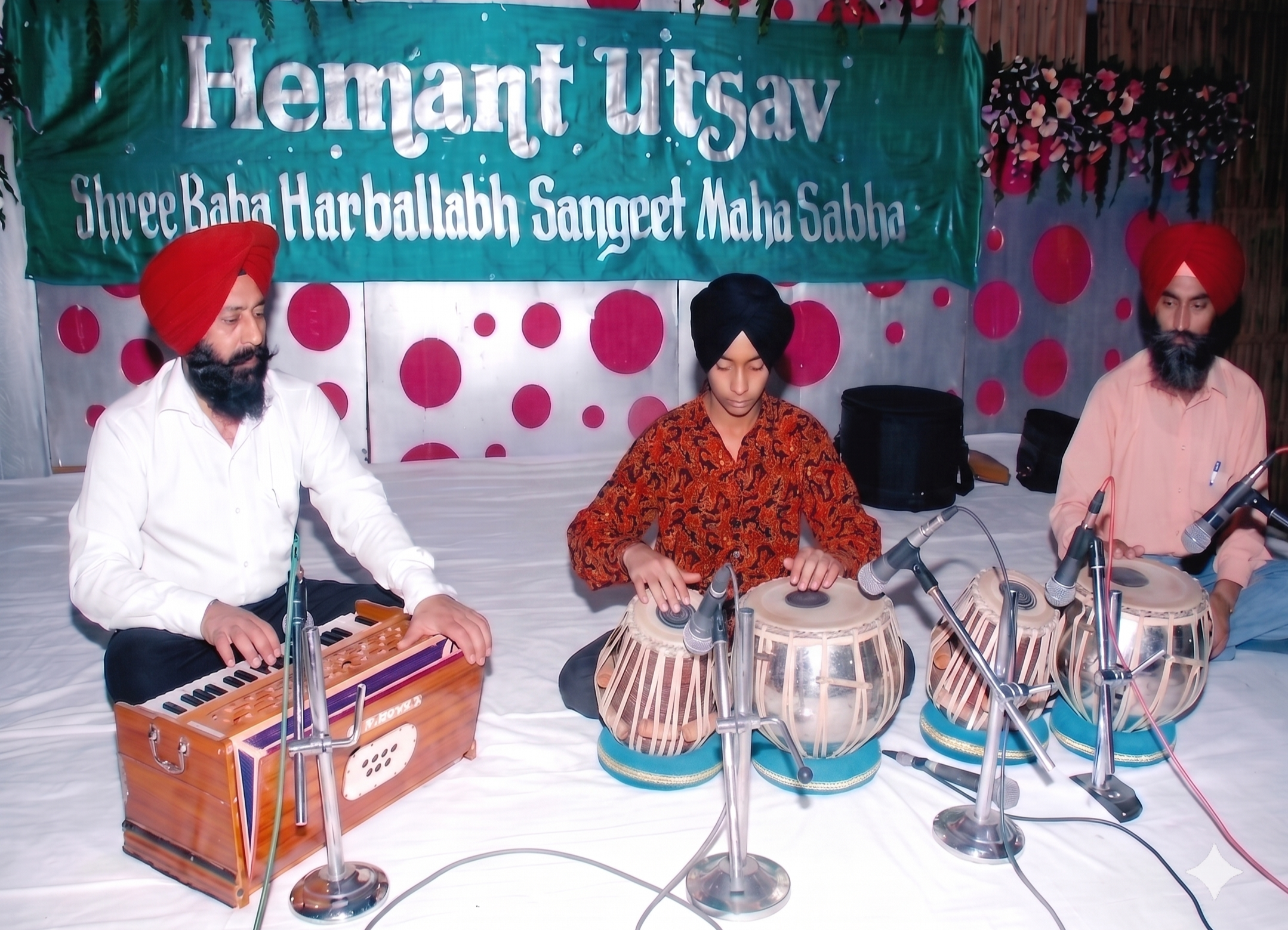 Three Indian musicians playing traditional instruments at a cultural event, with a green banner in the background.