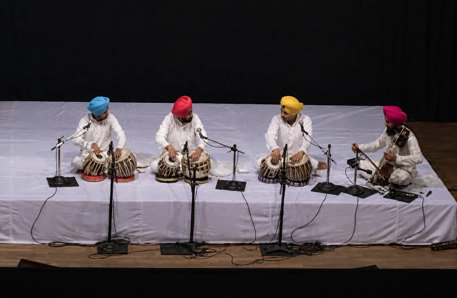 Four men dressed in white traditional attire and turbans, sitting on a stage with white cloth, playing Indian classical instruments: two tablas, a harmonium, and a sitar.