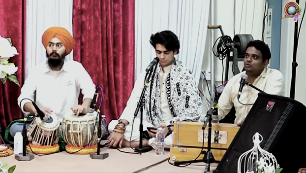 Three young men sitting at a table with microphones, musical instruments, and a water bottle during a performance or discussion in a room with red and white curtains.