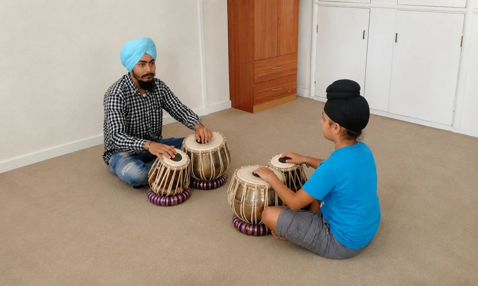 Two people sitting on the floor playing traditional Indian drums, one man with a turban and the other with a head wrap.