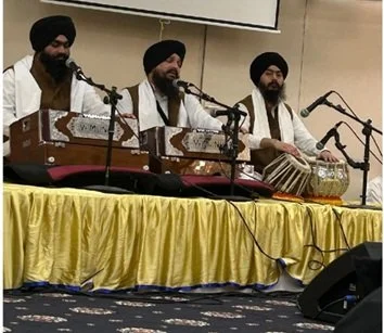 Three musicians wearing turbans playing traditional Indian musical instruments on a stage with a yellow cloth skirt.