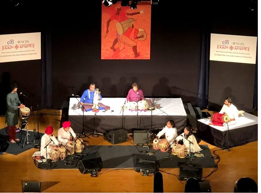 Musicians performing on stage, playing traditional Indian instruments, with a large painting of a classical dance scene hanging above them.