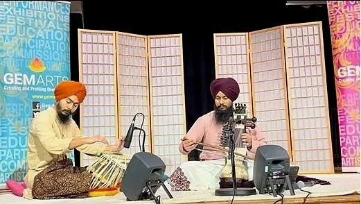 Two men wearing traditional turbans seated on stage with musical instruments, microphones, and speakers, behind folding screens with colorful banners.