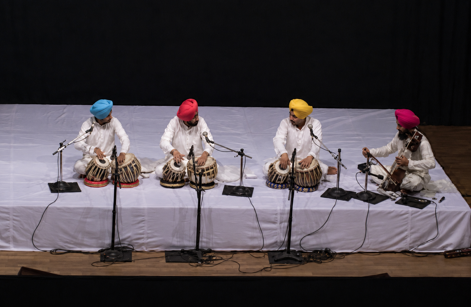 Five men wearing white traditional attire and colorful turbans playing classical Indian music on stage. Four are playing tabla drums and one is playing a sitar, with microphones in front of them.