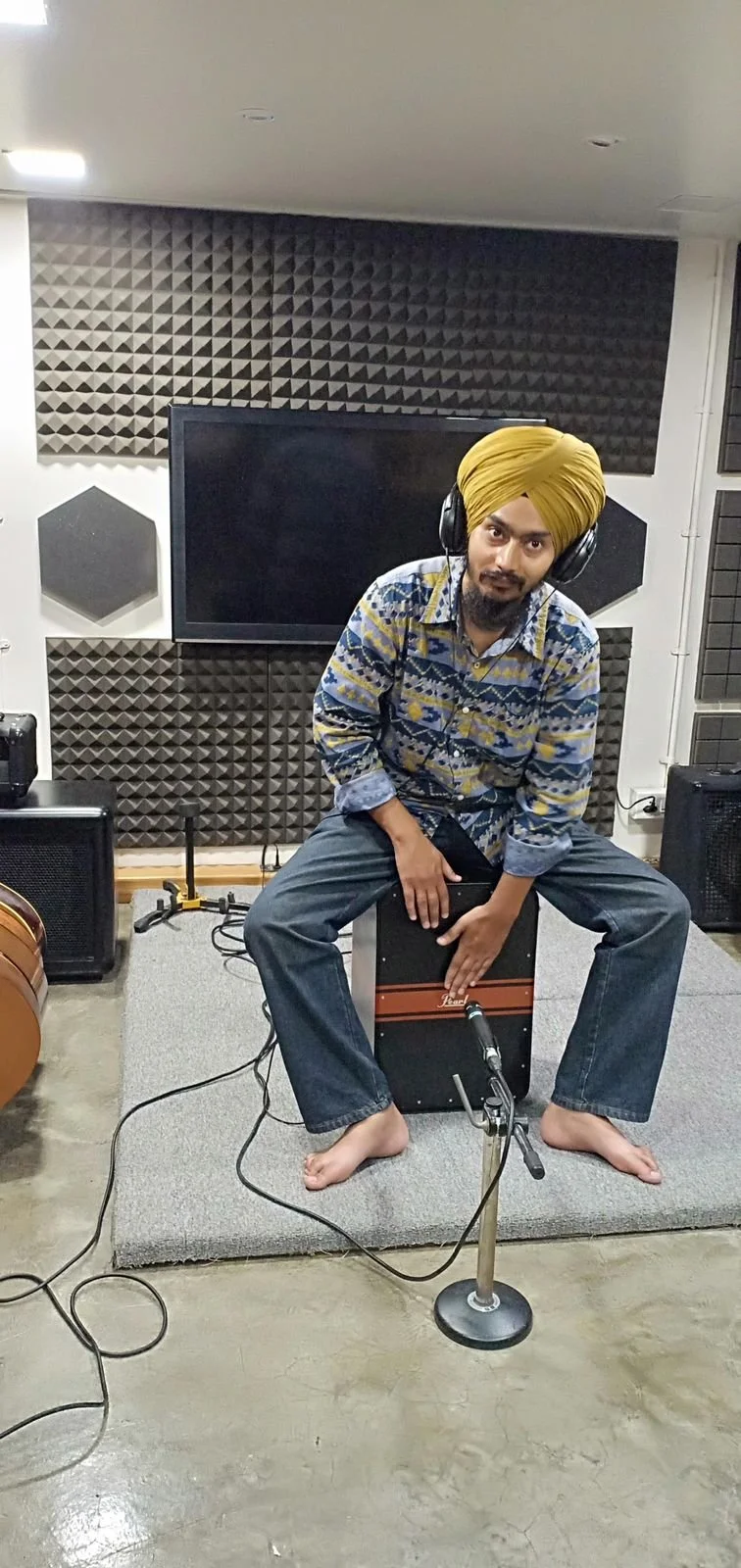 A man with a yellow turban and patterned shirt sitting on a cajón drum in a recording studio with soundproofing foam, microphone, and audio equipment.