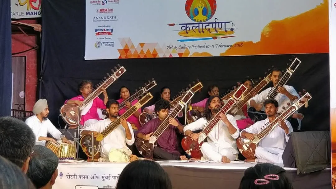 Group of musicians performing on stage with sitar and tabla instruments during an art and culture festival in India, with festival banner in the background.