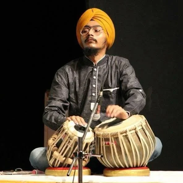 A man wearing a yellow turban, glasses, and a dark traditional kurta playing a pair of tabla drums on stage.