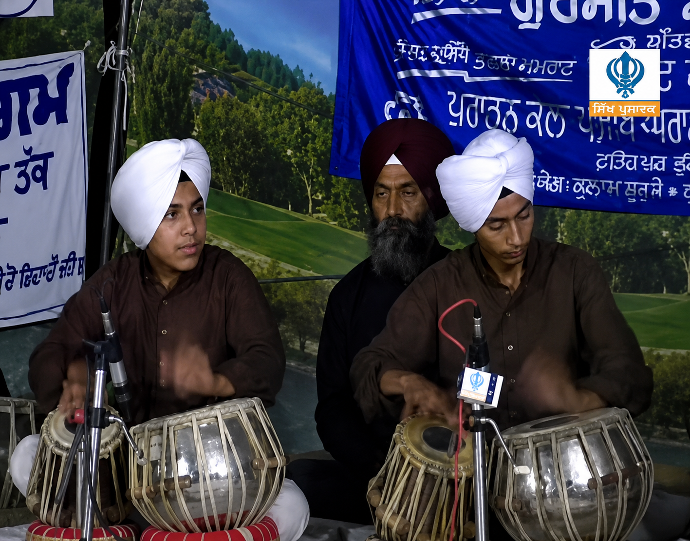 Three men in traditional Sikh attire playing musical instruments called tablas during a cultural event. They are seated on the stage with a backdrop featuring lush green landscape and blue text banners.