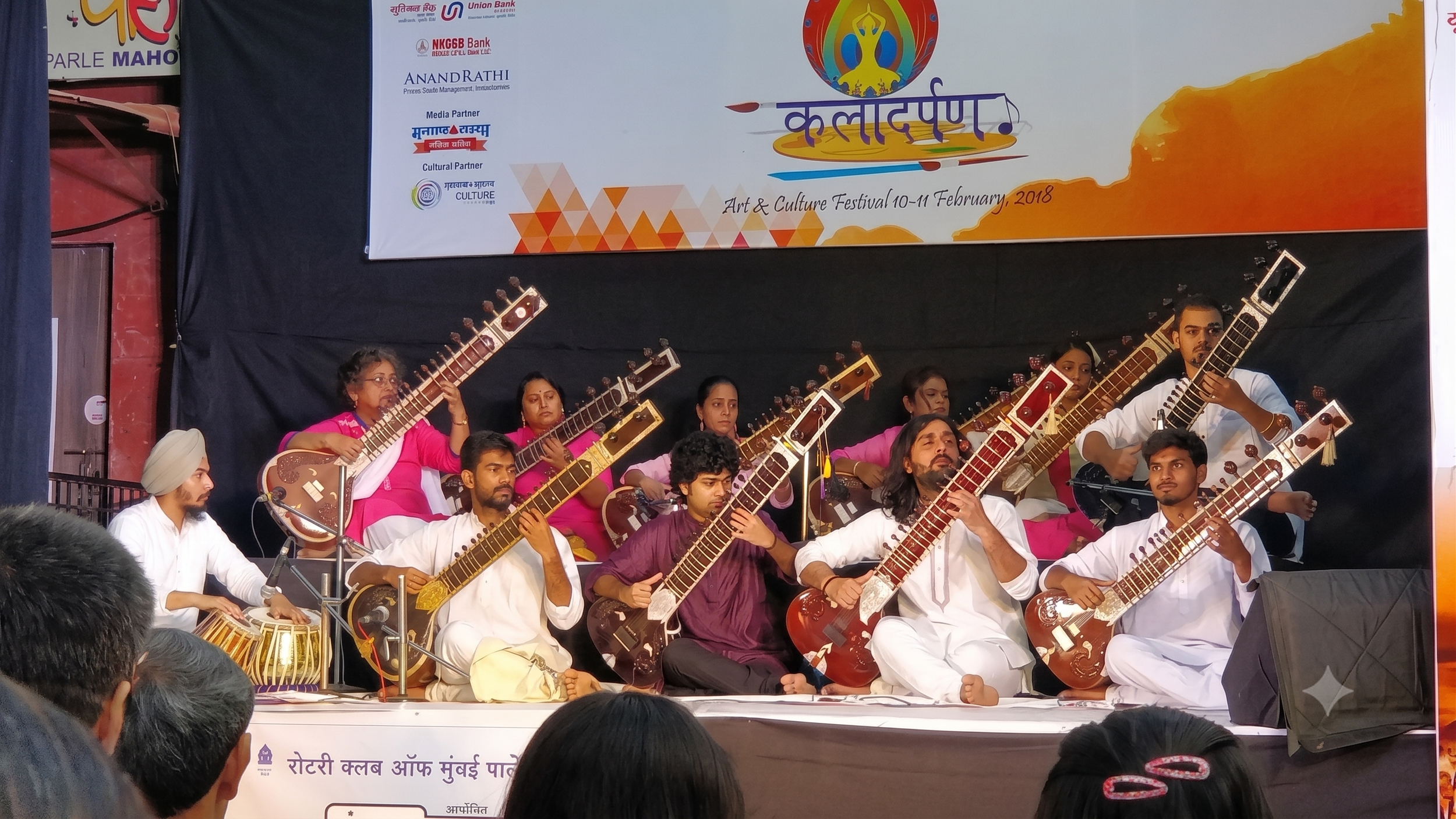 Group of musicians performing on stage at an art and culture festival, with a large banner above them displaying event details and sponsors.