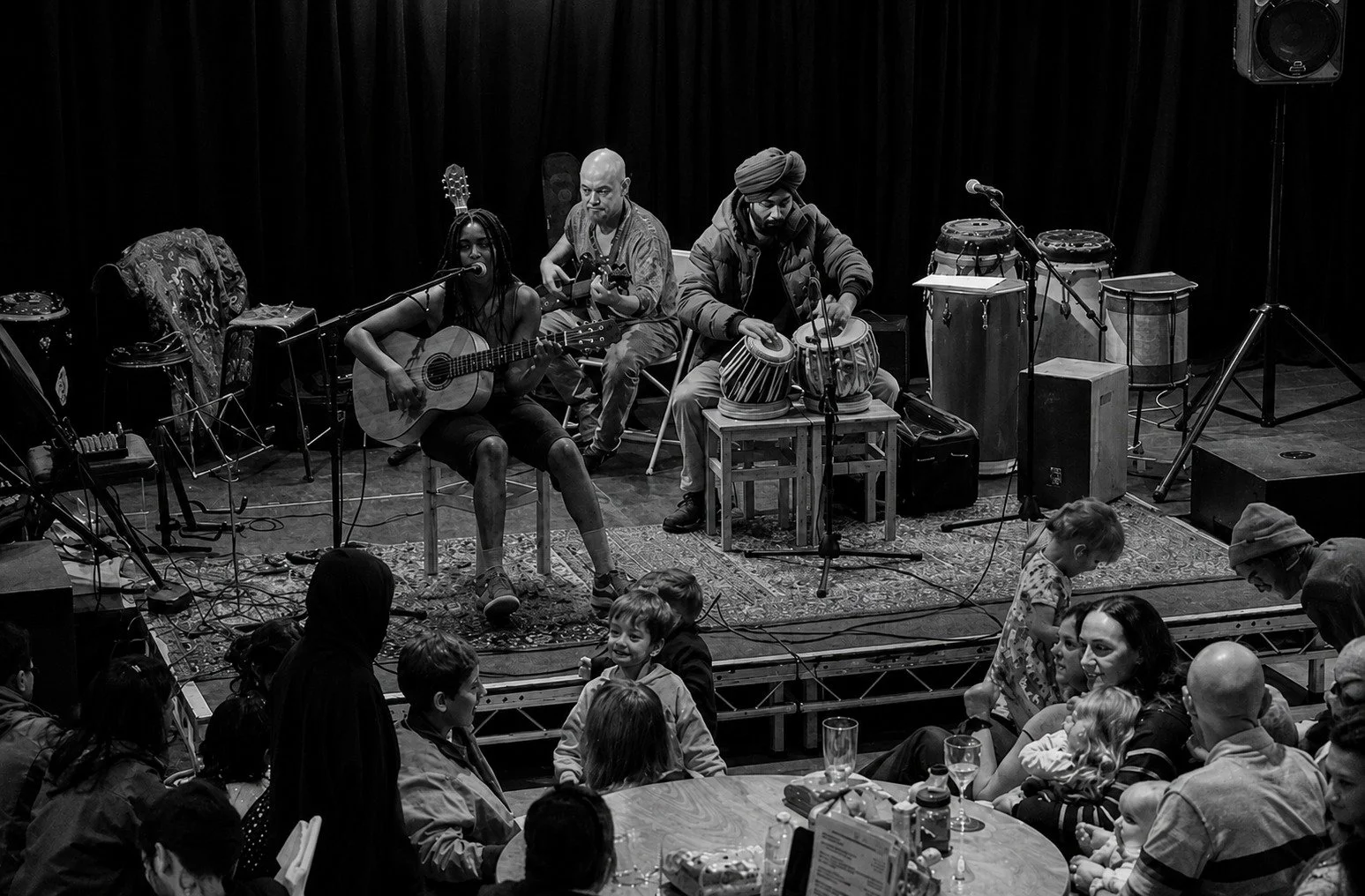 A black-and-white photo of a musical performance on stage with three musicians playing instruments and a group of children and adults sitting and standing in front of the stage, enjoying the show.
